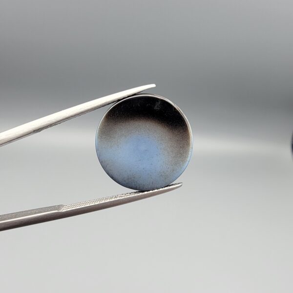 Multi-angle macro photographs of a 12.87-carat natural African Banded Eye Agate (Sulemani Aqeeq) held in gem tweezers, displaying a high round cabochon dome with concentric white and grey fortification rings, a brownish-black core pupil, and a base showing soft blue-grey to dark gradient chalcedony.