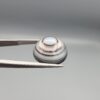 Multi-angle macro photographs of a 12.76-carat natural African Banded Eye Agate (Sulemani Aqeeq) held in gem tweezers, displaying a high round cabochon dome with concentric white and dark brown fortification rings framing a defined eye motif, tiered side profile banding, and a smooth dark brown base.