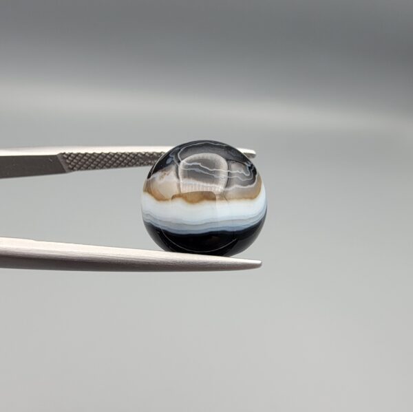 Multi-angle macro photographs of an 8.02-carat natural African Banded Eye Agate (Sulemani Aqeeq) held in steel gem tweezers, displaying a high round cabochon dome with wavy concentric white and earth-brown banding around a deep black core, crisp side fortification layers, and a flat jet-black base.
