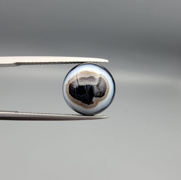 Multi-angle macro photographs of a 6.94-carat natural African Banded Eye Agate (Sulemani Aqeeq) held in gem tweezers, displaying a high round cabochon dome with complex concentric white and brown fortification rings around a deep black core, tiered profile layers, and a polished flat black base.