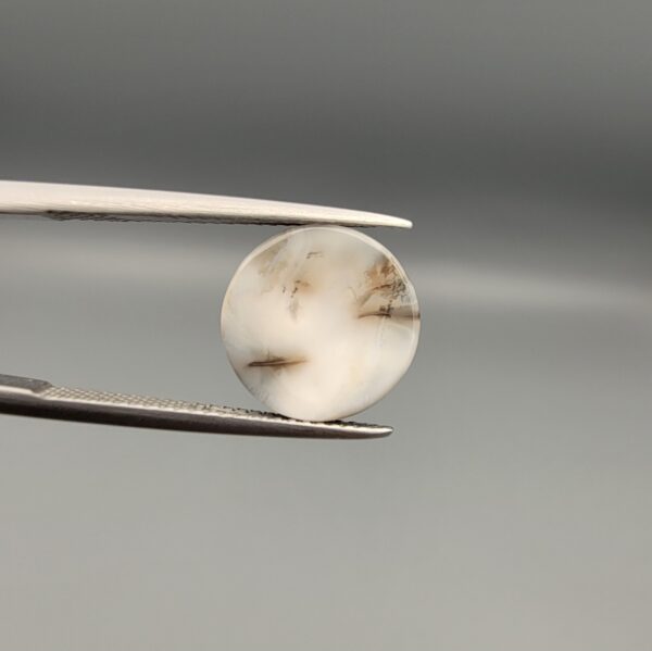 Multi-angle macro photographs of a 5.62-carat natural African Banded Eye Agate (Sulemani Aqeeq) held in steel gem tweezers, showcasing a high round cabochon dome with bold concentric white and jet-black banding framing a brownish-black pupil core, crisp side profile layers, and a flat white base with natural dark dendrite plumes.