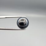 Multi-angle macro photographs of a 6.67-carat natural African Banded Eye Agate (Sulemani Aqeeq) held in gem tweezers, showing a high round cabochon dome with a deep onyx-black body framed by a sharp, concentric white outer arc, smooth profile curvature, and a polished solid black reverse base.