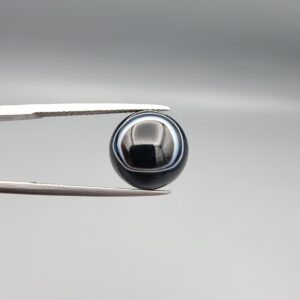 Multi-angle macro photographs of a 6.67-carat natural African Banded Eye Agate (Sulemani Aqeeq) held in gem tweezers, showing a high round cabochon dome with a deep onyx-black body framed by a sharp, concentric white outer arc, smooth profile curvature, and a polished solid black reverse base.