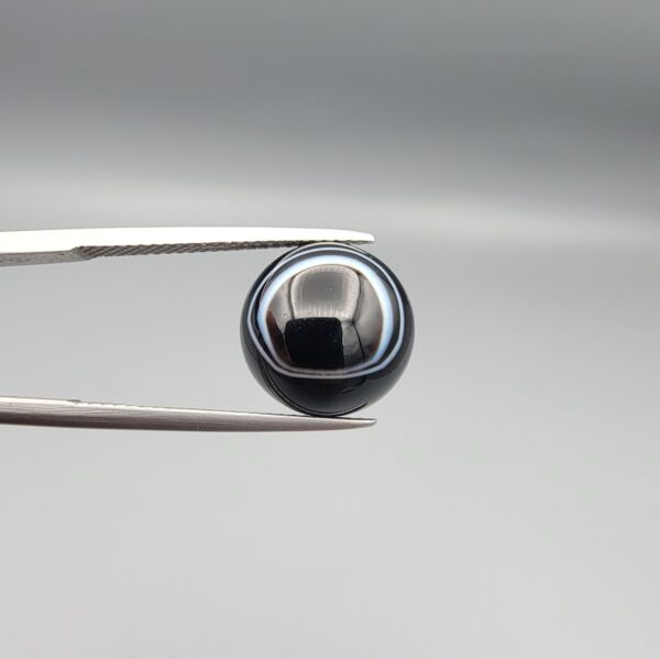 Multi-angle macro photographs of a 6.67-carat natural African Banded Eye Agate (Sulemani Aqeeq) held in gem tweezers, showing a high round cabochon dome with a deep onyx-black body framed by a sharp, concentric white outer arc, smooth profile curvature, and a polished solid black reverse base.