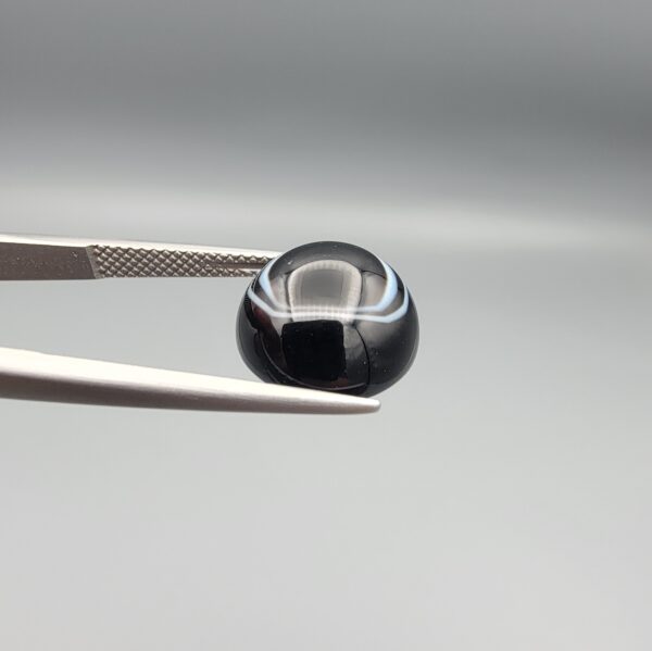 Multi-angle macro photographs of a 6.67-carat natural African Banded Eye Agate (Sulemani Aqeeq) held in gem tweezers, showing a high round cabochon dome with a deep onyx-black body framed by a sharp, concentric white outer arc, smooth profile curvature, and a polished solid black reverse base.