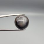 Multi-angle macro photographs of a 13.29-carat natural African Banded Eye Agate (Sulemani Aqeeq) shown in gem tweezers and held on a finger, featuring a high round cabochon dome with a deep charcoal body, subtle translucent inner zoning, layered side fortification bands, and a smooth black base.