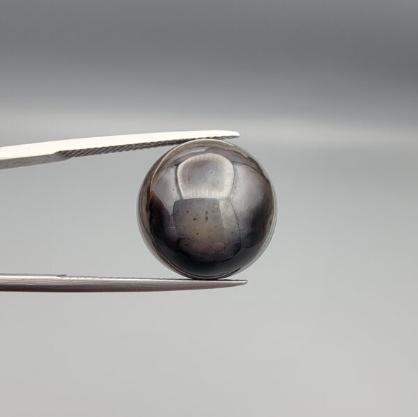 Multi-angle macro photographs of a 13.29-carat natural African Banded Eye Agate (Sulemani Aqeeq) shown in gem tweezers and held on a finger, featuring a high round cabochon dome with a deep charcoal body, subtle translucent inner zoning, layered side fortification bands, and a smooth black base.