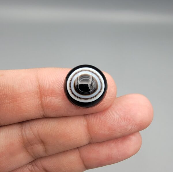 Multi-angle macro photographs of a 14.55-carat natural African Banded Eye Agate (Sulemani Aqeeq) held on fingers, showcasing a high round cabochon dome with sharp concentric white and black fortification rings framing a deep black pupil core, and a smooth jet-black base.