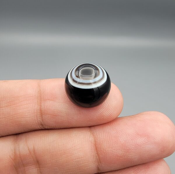 Multi-angle macro photographs of a 14.55-carat natural African Banded Eye Agate (Sulemani Aqeeq) held on fingers, showcasing a high round cabochon dome with sharp concentric white and black fortification rings framing a deep black pupil core, and a smooth jet-black base.