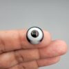Multi-angle macro photographs of a 12.40-carat natural African Banded Eye Agate (Sulemani Aqeeq) held on fingertips, displaying a high round cabochon dome with a prominent central black pupil enclosed by a bold white ring, smooth black outer border, and a flat reverse showing a dark smoky-grey gradient base.
