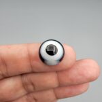 Multi-angle macro photographs of a 12.40-carat natural African Banded Eye Agate (Sulemani Aqeeq) held on fingertips, displaying a high round cabochon dome with a prominent central black pupil enclosed by a bold white ring, smooth black outer border, and a flat reverse showing a dark smoky-grey gradient base.