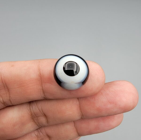 Multi-angle macro photographs of a 12.40-carat natural African Banded Eye Agate (Sulemani Aqeeq) held on fingertips, displaying a high round cabochon dome with a prominent central black pupil enclosed by a bold white ring, smooth black outer border, and a flat reverse showing a dark smoky-grey gradient base.