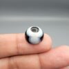 Multi-angle macro photographs of a 12.40-carat natural African Banded Eye Agate (Sulemani Aqeeq) held on fingertips, displaying a high round cabochon dome with a prominent central black pupil enclosed by a bold white ring, smooth black outer border, and a flat reverse showing a dark smoky-grey gradient base.