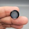 Multi-angle macro photographs of a 12.40-carat natural African Banded Eye Agate (Sulemani Aqeeq) held on fingertips, displaying a high round cabochon dome with a prominent central black pupil enclosed by a bold white ring, smooth black outer border, and a flat reverse showing a dark smoky-grey gradient base.