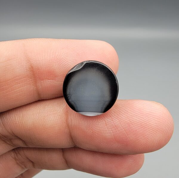 Multi-angle macro photographs of a 12.40-carat natural African Banded Eye Agate (Sulemani Aqeeq) held on fingertips, displaying a high round cabochon dome with a prominent central black pupil enclosed by a bold white ring, smooth black outer border, and a flat reverse showing a dark smoky-grey gradient base.