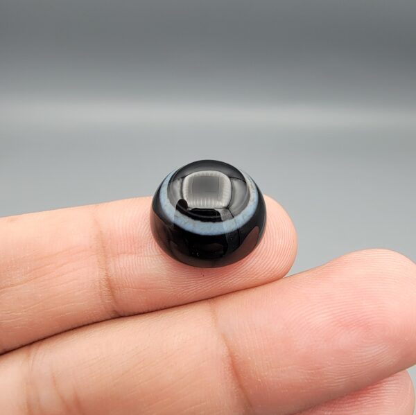 Multi-angle macro photographs of a 14.09-carat natural African Banded Eye Agate (Sulemani Aqeeq) held on fingertips, displaying a high round cabochon dome with a deep onyx-black core framed by sharp concentric white outer ringing, layered side profile banding, and a smooth jet-black reverse base.