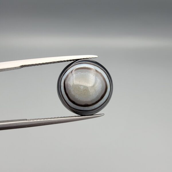 Multi-angle macro photographs of a 12.57-carat natural African Banded Eye Agate (Sulemani Aqeeq) held in gem tweezers and on fingertips, showcasing a high round cabochon dome with concentric white and dark grey bands framing a luminous translucent central dome, layered profile fortification rings, and a flat reverse showing an organic dark brown and black cloud matrix.