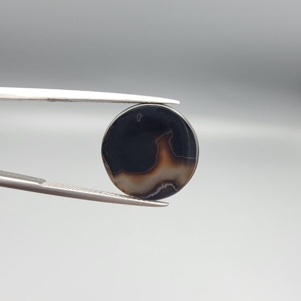 Multi-angle macro photographs of a 12.57-carat natural African Banded Eye Agate (Sulemani Aqeeq) held in gem tweezers and on fingertips, showcasing a high round cabochon dome with concentric white and dark grey bands framing a luminous translucent central dome, layered profile fortification rings, and a flat reverse showing an organic dark brown and black cloud matrix.