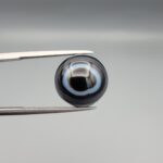 Multi-angle macro photographs of a 13.11-carat natural African Banded Eye Agate (Sulemani Aqeeq) held in gem tweezers, displaying a high round cabochon dome with crisp concentric white fortification rings around a deep black pupil core, dramatic side profile banding, and a reverse base showcasing a bold white-and-grey crescent wave pattern.