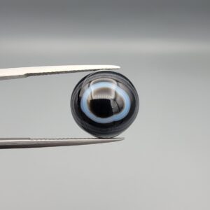 Multi-angle macro photographs of a 13.11-carat natural African Banded Eye Agate (Sulemani Aqeeq) held in gem tweezers, displaying a high round cabochon dome with crisp concentric white fortification rings around a deep black pupil core, dramatic side profile banding, and a reverse base showcasing a bold white-and-grey crescent wave pattern.
