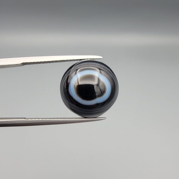 Multi-angle macro photographs of a 13.11-carat natural African Banded Eye Agate (Sulemani Aqeeq) held in gem tweezers, displaying a high round cabochon dome with crisp concentric white fortification rings around a deep black pupil core, dramatic side profile banding, and a reverse base showcasing a bold white-and-grey crescent wave pattern.