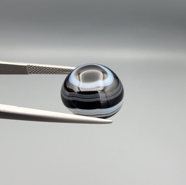 Multi-angle macro photographs of a 13.11-carat natural African Banded Eye Agate (Sulemani Aqeeq) held in gem tweezers, displaying a high round cabochon dome with crisp concentric white fortification rings around a deep black pupil core, dramatic side profile banding, and a reverse base showcasing a bold white-and-grey crescent wave pattern.