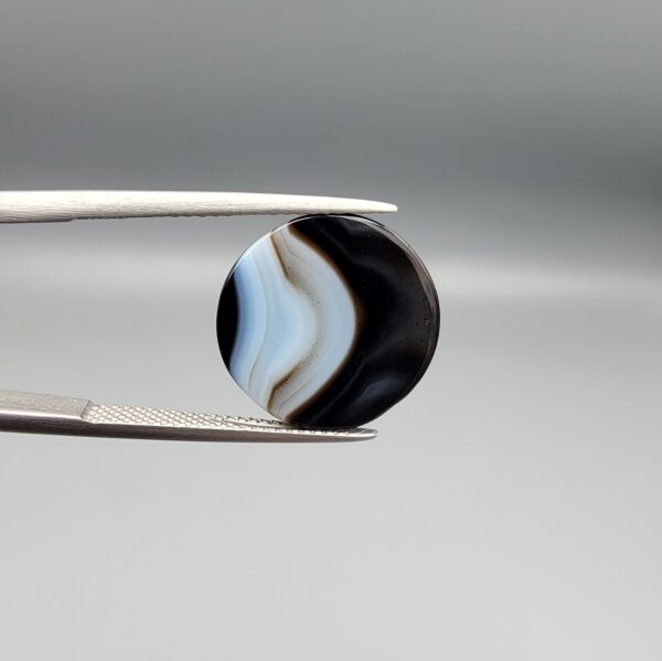 Multi-angle macro photographs of a 13.11-carat natural African Banded Eye Agate (Sulemani Aqeeq) held in gem tweezers, displaying a high round cabochon dome with crisp concentric white fortification rings around a deep black pupil core, dramatic side profile banding, and a reverse base showcasing a bold white-and-grey crescent wave pattern.