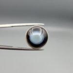 Multi-angle macro photographs of a 10.42-carat natural African Banded Eye Agate (Sulemani Aqeeq) held in tweezers and on fingertips, displaying a high round cabochon dome with a luminous translucent ice-blue eye core framed by dark fortification rings, layered side flank banding, and a webbed blue-grey chalcedony base.