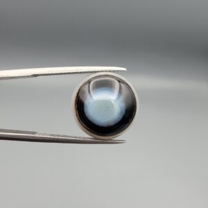 Multi-angle macro photographs of a 10.42-carat natural African Banded Eye Agate (Sulemani Aqeeq) held in tweezers and on fingertips, displaying a high round cabochon dome with a luminous translucent ice-blue eye core framed by dark fortification rings, layered side flank banding, and a webbed blue-grey chalcedony base.