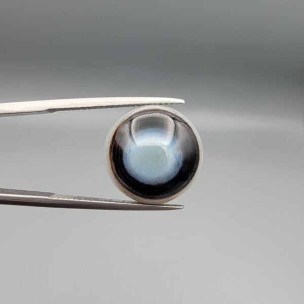 Multi-angle macro photographs of a 10.42-carat natural African Banded Eye Agate (Sulemani Aqeeq) held in tweezers and on fingertips, displaying a high round cabochon dome with a luminous translucent ice-blue eye core framed by dark fortification rings, layered side flank banding, and a webbed blue-grey chalcedony base.