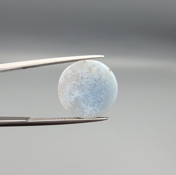 Multi-angle macro photographs of a 10.42-carat natural African Banded Eye Agate (Sulemani Aqeeq) held in tweezers and on fingertips, displaying a high round cabochon dome with a luminous translucent ice-blue eye core framed by dark fortification rings, layered side flank banding, and a webbed blue-grey chalcedony base.