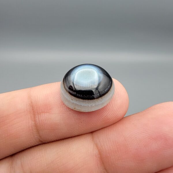 Multi-angle macro photographs of a 10.42-carat natural African Banded Eye Agate (Sulemani Aqeeq) held in tweezers and on fingertips, displaying a high round cabochon dome with a luminous translucent ice-blue eye core framed by dark fortification rings, layered side flank banding, and a webbed blue-grey chalcedony base.
