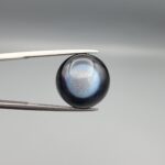 Macro photographs of an 11.69-carat natural African Banded Eye Agate (Sulemani Aqeeq) held in steel gem tweezers, displaying a high round cabochon dome with a deep onyx-black body centered by a luminous blue-grey eye motif, and a polished flat black base.