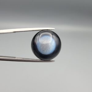 Macro photographs of an 11.69-carat natural African Banded Eye Agate (Sulemani Aqeeq) held in steel gem tweezers, displaying a high round cabochon dome with a deep onyx-black body centered by a luminous blue-grey eye motif, and a polished flat black base.