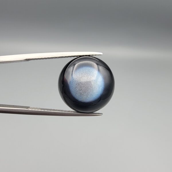 Macro photographs of an 11.69-carat natural African Banded Eye Agate (Sulemani Aqeeq) held in steel gem tweezers, displaying a high round cabochon dome with a deep onyx-black body centered by a luminous blue-grey eye motif, and a polished flat black base.