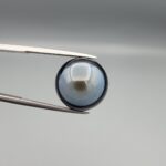 Multi-angle macro photographs of a 13.83-carat natural African Banded Eye Agate (Sulemani Aqeeq) held in steel gem tweezers, showcasing a high round cabochon dome with a luminous translucent ice-blue central eye field, framed by a deep black margin, and a smooth jet-black flat reverse base.