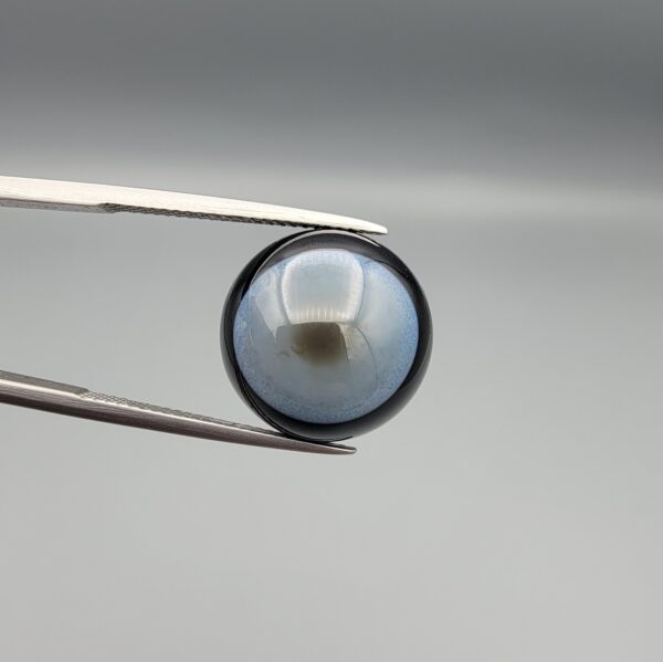 Multi-angle macro photographs of a 13.83-carat natural African Banded Eye Agate (Sulemani Aqeeq) held in steel gem tweezers, showcasing a high round cabochon dome with a luminous translucent ice-blue central eye field, framed by a deep black margin, and a smooth jet-black flat reverse base.
