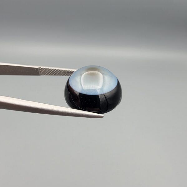Multi-angle macro photographs of a 13.83-carat natural African Banded Eye Agate (Sulemani Aqeeq) held in steel gem tweezers, showcasing a high round cabochon dome with a luminous translucent ice-blue central eye field, framed by a deep black margin, and a smooth jet-black flat reverse base.