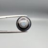 Multi-angle macro photographs of a 10.47-carat natural African Banded Eye Agate (Sulemani Aqeeq) shown in gem tweezers and on fingertips, featuring a high round cabochon dome with concentric black, brown, and white fortification rings framing a luminous pale blue-grey eye core, parallel layered profile banding, and a dark mottled reverse base with a nebulous center.