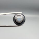 Multi-angle macro photographs of a 10.47-carat natural African Banded Eye Agate (Sulemani Aqeeq) shown in gem tweezers and on fingertips, featuring a high round cabochon dome with concentric black, brown, and white fortification rings framing a luminous pale blue-grey eye core, parallel layered profile banding, and a dark mottled reverse base with a nebulous center.