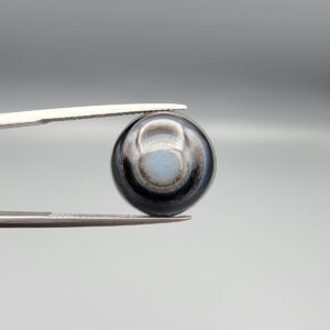 Multi-angle macro photographs of a 10.47-carat natural African Banded Eye Agate (Sulemani Aqeeq) shown in gem tweezers and on fingertips, featuring a high round cabochon dome with concentric black, brown, and white fortification rings framing a luminous pale blue-grey eye core, parallel layered profile banding, and a dark mottled reverse base with a nebulous center.