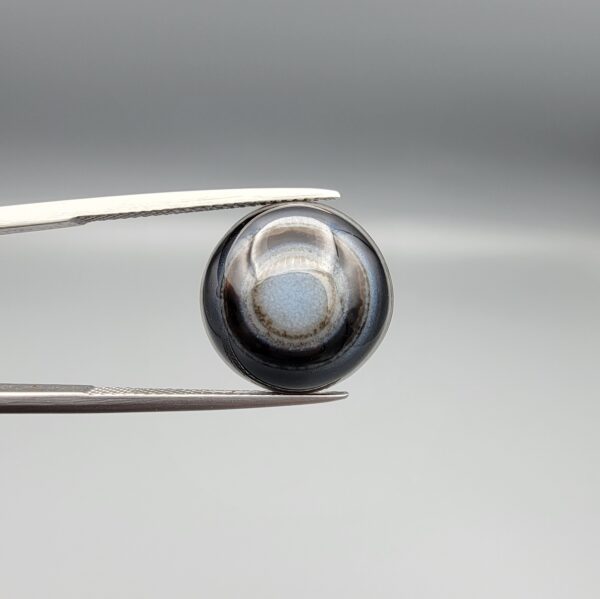 Multi-angle macro photographs of a 10.47-carat natural African Banded Eye Agate (Sulemani Aqeeq) shown in gem tweezers and on fingertips, featuring a high round cabochon dome with concentric black, brown, and white fortification rings framing a luminous pale blue-grey eye core, parallel layered profile banding, and a dark mottled reverse base with a nebulous center.