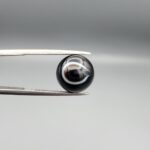 Multi-angle macro photographs of a 7.14-carat natural African Banded Eye Agate (Sulemani Aqeeq) shown in gem tweezers and on fingertips, featuring a high round cabochon dome with a deep black body accented by a sharp teardrop-shaped concentric white and black ring pattern forming a distinct central eye motif, and a solid uniform black reverse base with minor edge wear.