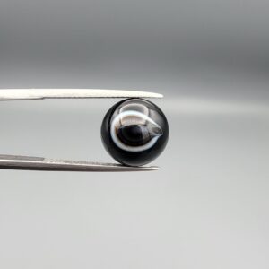 Multi-angle macro photographs of a 7.14-carat natural African Banded Eye Agate (Sulemani Aqeeq) shown in gem tweezers and on fingertips, featuring a high round cabochon dome with a deep black body accented by a sharp teardrop-shaped concentric white and black ring pattern forming a distinct central eye motif, and a solid uniform black reverse base with minor edge wear.