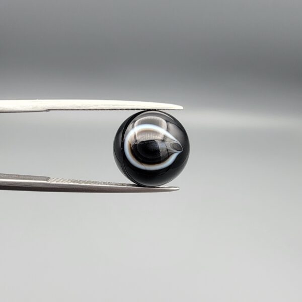 Multi-angle macro photographs of a 7.14-carat natural African Banded Eye Agate (Sulemani Aqeeq) shown in gem tweezers and on fingertips, featuring a high round cabochon dome with a deep black body accented by a sharp teardrop-shaped concentric white and black ring pattern forming a distinct central eye motif, and a solid uniform black reverse base with minor edge wear.