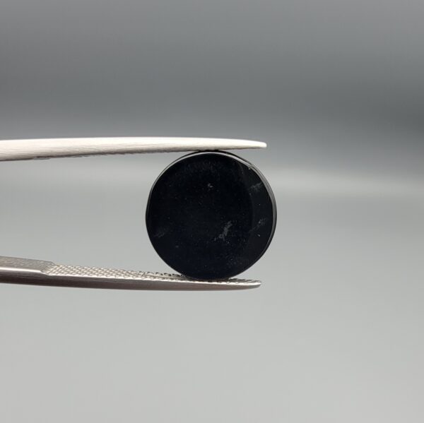 Multi-angle macro photographs of a 7.14-carat natural African Banded Eye Agate (Sulemani Aqeeq) shown in gem tweezers and on fingertips, featuring a high round cabochon dome with a deep black body accented by a sharp teardrop-shaped concentric white and black ring pattern forming a distinct central eye motif, and a solid uniform black reverse base with minor edge wear.