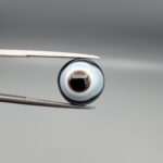 Multi-angle macro photographs of a 9.85-carat natural African Banded Eye Agate (Sulemani Aqeeq) held in gem tweezers, featuring a top view with a central eye motif of concentric white and dark rings set in a deep black body, a profile view demonstrating smooth high-dome geometry and layered fortification banding, and a reverse view of a flat solid black base.