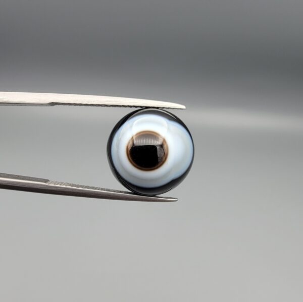 Multi-angle macro photographs of a 9.85-carat natural African Banded Eye Agate (Sulemani Aqeeq) held in gem tweezers, featuring a top view with a central eye motif of concentric white and dark rings set in a deep black body, a profile view demonstrating smooth high-dome geometry and layered fortification banding, and a reverse view of a flat solid black base.