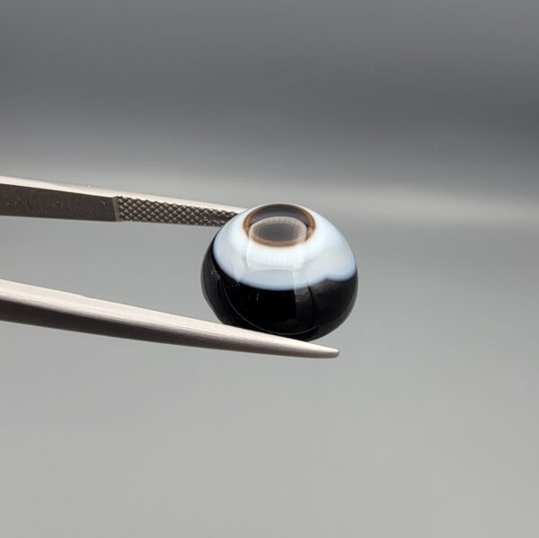 Multi-angle macro photographs of a 9.85-carat natural African Banded Eye Agate (Sulemani Aqeeq) held in gem tweezers, featuring a top view with a central eye motif of concentric white and dark rings set in a deep black body, a profile view demonstrating smooth high-dome geometry and layered fortification banding, and a reverse view of a flat solid black base.