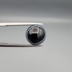 Multi-angle macro photographs of a 5.97-carat natural African Banded Eye Agate (Sulemani Aqeeq) held in gem tweezers, showing a top view with a dominant black body framed by a sharp white arc, a profile view with a distinct V-shaped white fortification band, and a reverse view revealing a multi-eye cellular pattern in blue-grey and black.